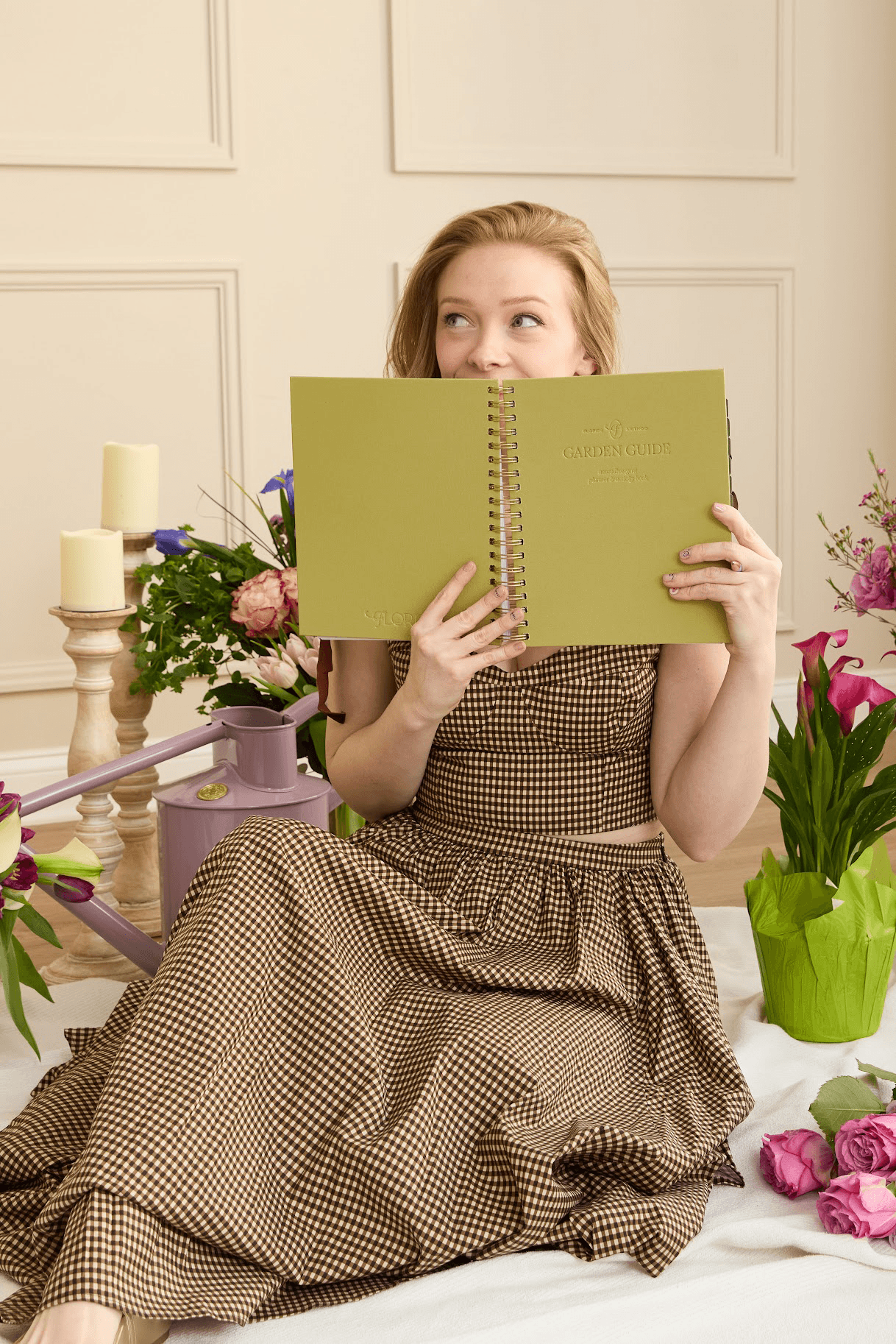 Woman in a checkered dress holding a green Floriss Garden Guide planner, playfully hiding her face, seated among fresh flowers and candles in a cozy, garden-inspired space representing The Grove community