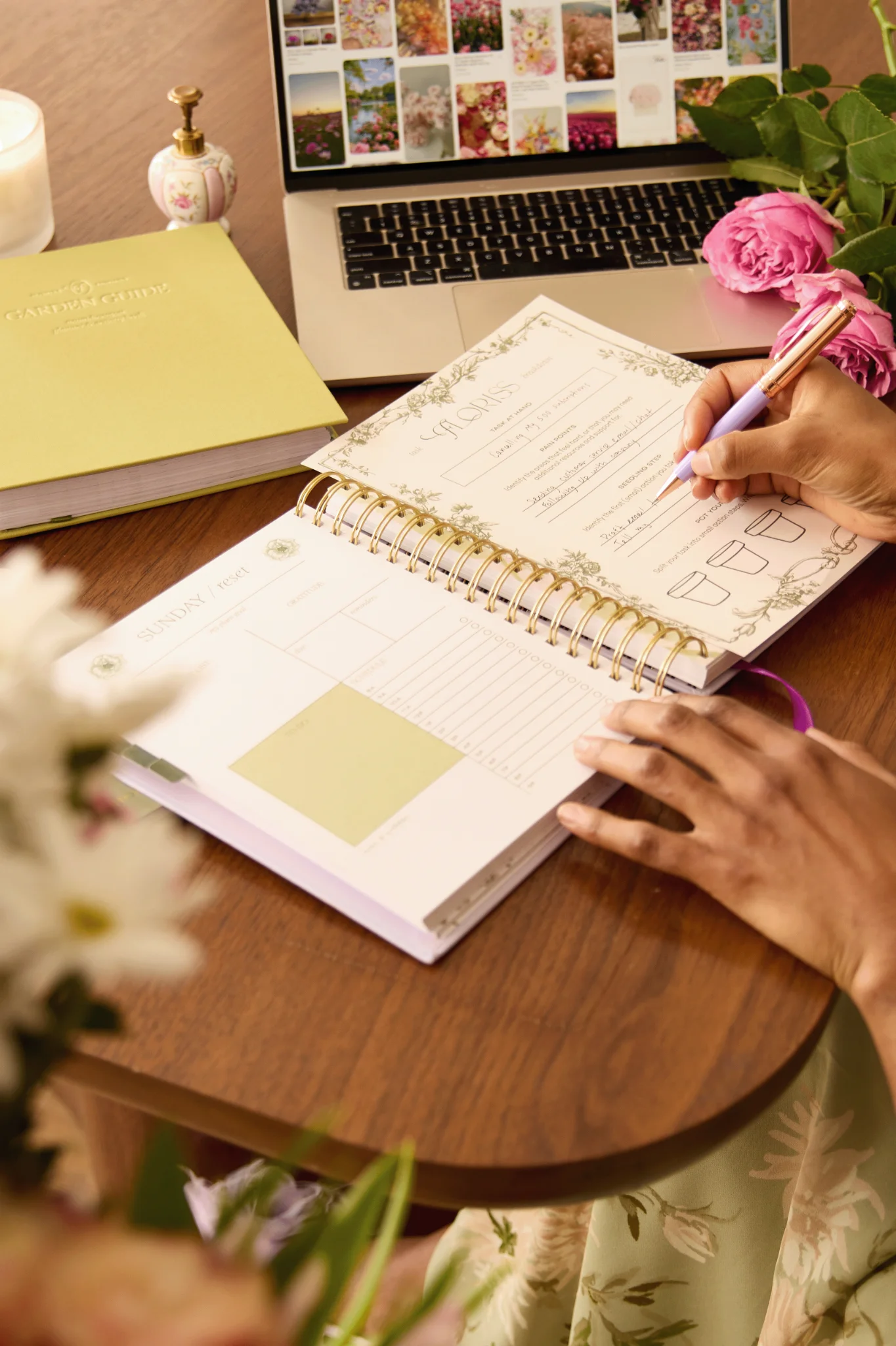 A woman writes in the Floriss Garden Guide planner on a warm wooden table, surrounded by fresh roses and a laptop open to a floral mood board representing Floriss' Garden Clubs community platform. The setting is quietly intentional — a moment of rhythm, beauty, and personal bloom.